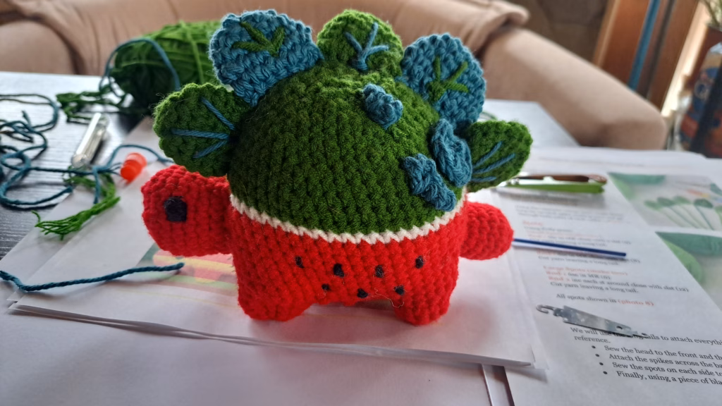 Ein entzückender, selbstgehäkelter Plüsch-Saurier mit einem grünen, blattartigen Körper und einem roten Unterteil, der auf einem Tisch steht.