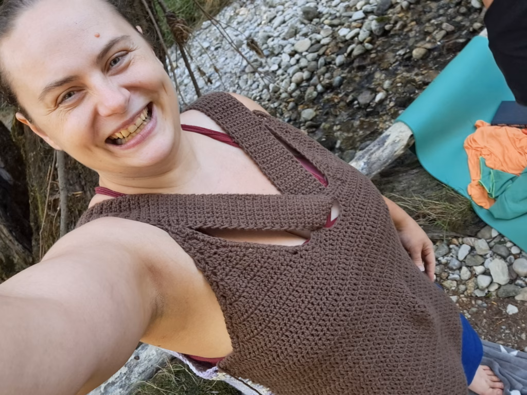 Ein fröhliches Selfie einer Frau, die ein selbstgehäkeltes oder gestricktes braunes Oberteil trägt, aufgenommen im Freien vor einem bewaldeten Hintergrund.