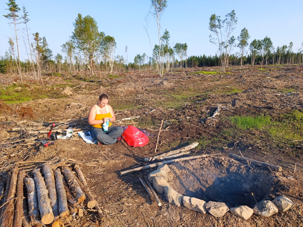 Eine Frau sitzt bei tiefstehender Sonne auf einer Rodungsfläche in Cape Breton und widmet sich ihrer Handarbeit, im Vordergrund eine rustikale Feuerstelle aus Steinen.